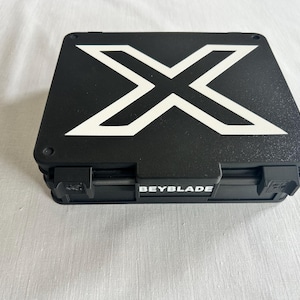 Può includere: Una custodia rettangolare nera con un grande design a "X" bianco sulla parte superiore. La parola "BEYBLADE" è stampata in bianco sulla parte anteriore della custodia. La custodia ha un meccanismo di chiusura sicuro.
