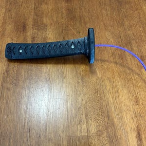 Op de afbeelding: Een zwart zwaardhandvat met een textuur van kruislings gearceerde patronen en twee zilverkleurige metalen klinknagels. Een lange, dunne, flexibele paarse plastic tie-wrap steekt uit het uiteinde van het handvat en buigt naar de rechterkant.