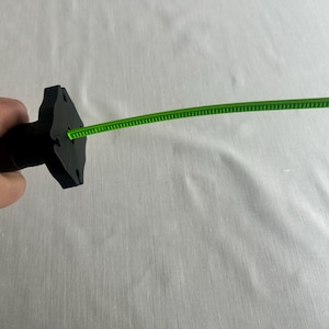 May include: A black object with a unique shape, possibly a handle, is held in a hand. A thin, bright green, flexible strip extends from the object. The background is a plain, light-colored surface.