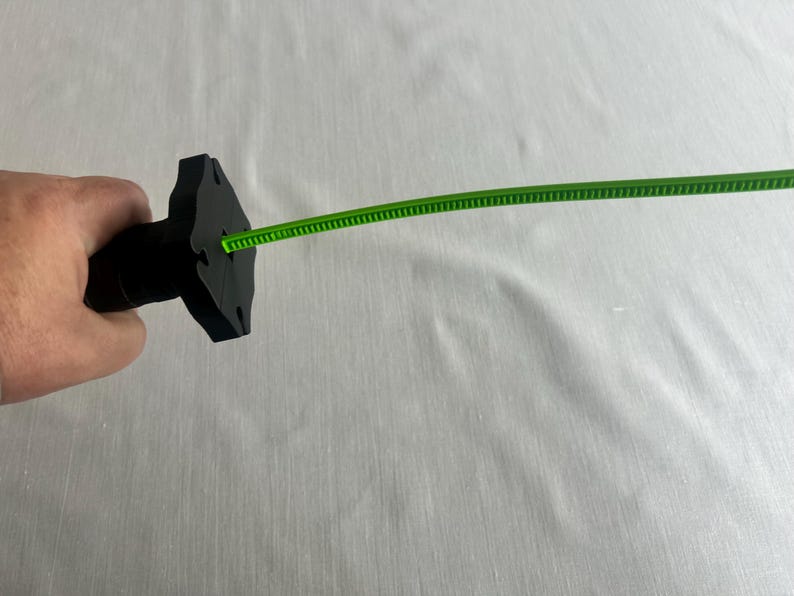 May include: A black object with a unique shape, possibly a handle, is held in a hand. A thin, bright green, flexible strip extends from the object. The background is a plain, light-colored surface.