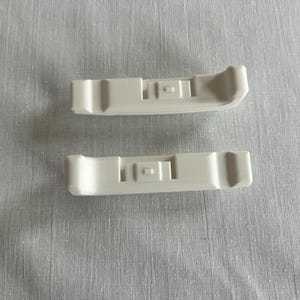 Op de afbeelding: Twee crèmekleurige plastic componenten, elk met een rechthoekig lichaam en afgeronde uiteinden. De componenten hebben een centraal verzonken gebied met twee kleine rechthoekige uitsparingen. De items zijn gerangschikt op een lichtgrijze stoffen ondergrond.