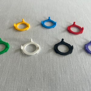 May include: Seven colorful, circular, plastic SIM card removal tools. The tools are in yellow, blue, red, green, white, black, and purple. Each tool has a ring shape with a small, pointed extension.
