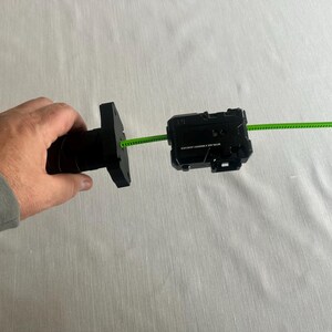 May include: A black toy launcher with a green pull string. The launcher is held in a person's hand. The launcher has a rectangular shape with a series of buttons and a small opening for the string.
