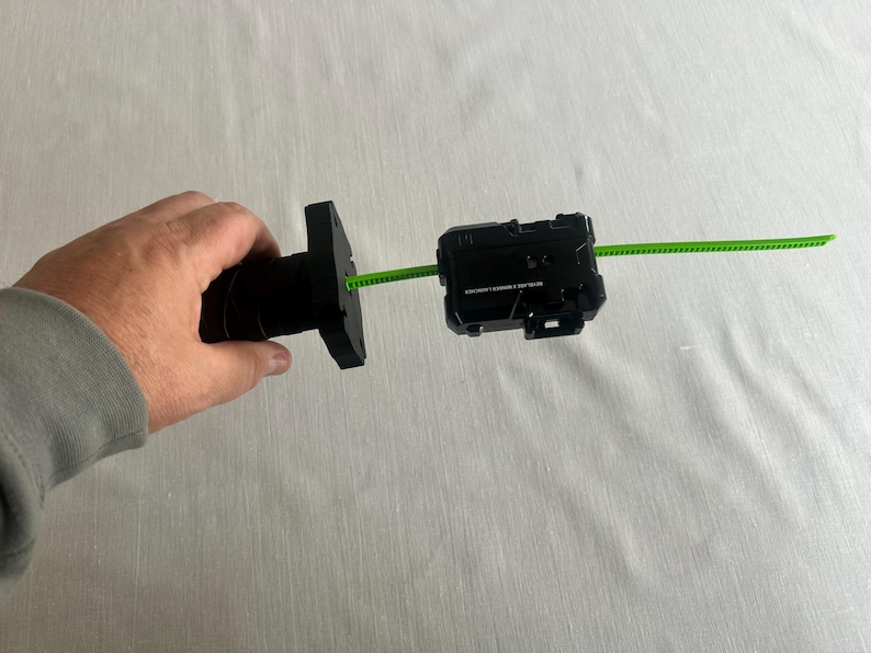 May include: A black toy launcher with a green pull string. The launcher is held in a person's hand. The launcher has a rectangular shape with a series of buttons and a small opening for the string.