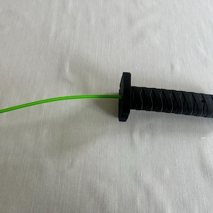 May include: A black handle with a textured grip and a square guard, with a bright green cord extending from the center. The handle has a cylindrical shape. The object is likely a component of a larger item.