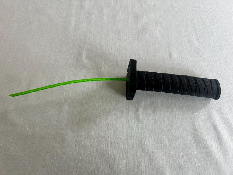 May include: A black handle with a textured grip and a square guard, with a bright green cord extending from the center. The handle has a cylindrical shape. The object is likely a component of a larger item.