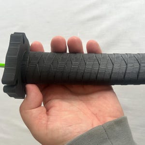 Puede incluir: Un puño de manillar de motocicleta negro con un patrón texturizado. El puño tiene forma cilíndrica con una brida en un extremo y un cable verde brillante que se extiende desde el otro extremo. El puño se sostiene en una mano.