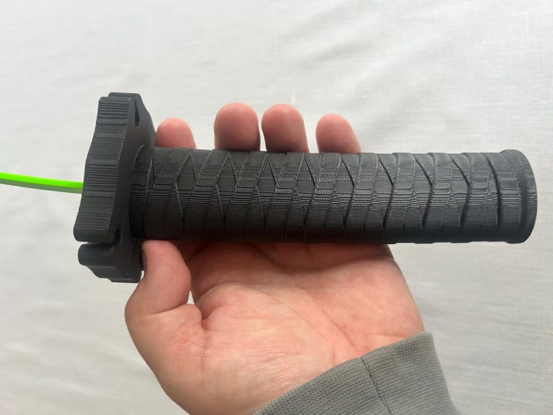 May include: A black motorcycle handlebar grip with a textured pattern. The grip has a cylindrical shape with a flange at one end and a bright green cable extending from the other end. The grip is held in a hand.