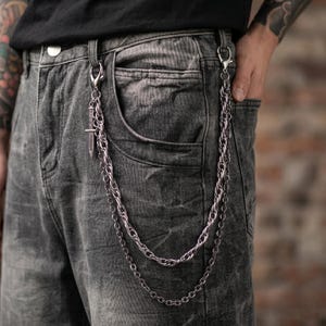 May include: A silver-colored, double-strand chain wallet accessory with a decorative cross pendant. The chain is attached to the belt loops and pocket of gray denim jeans. The jeans are worn with a black t-shirt.