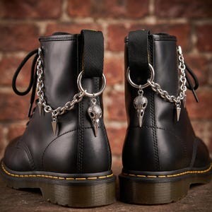 Peut inclure: Paire de bottes en cuir noir avec des chaînes argentées et des breloques en forme de crâne. Les bottes ont des coutures jaunes et des lacets noirs. Les breloques comprennent un pendentif crâne et des accents triangulaires argentés.