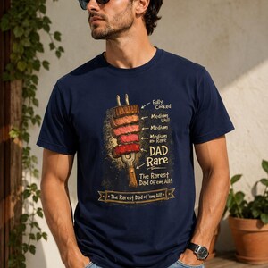 Könnte beinhalten: Ein marineblaues T-Shirt mit einer Grafik einer Gabel, die fünf Fleischstücke aufgespießt hat, welche mit Garstufen von Fully Cooked bis Rare beschriftet sind. Der Text lautet "DAD Rare" und "The Rarest Dad of 'em All!" auf einem Banner.