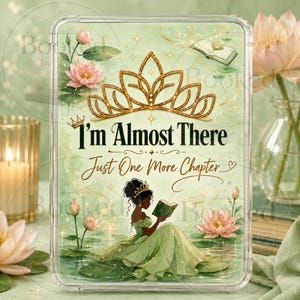 May include: A rectangular clear acrylic block featuring a printed illustration of a person in a green gown reading a book on a lily pad. The design includes a gold tiara, pink lotus flowers, and the text I'm Almost There Just One More Chapter.