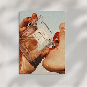 May include: A rectangular wall art print featuring a clear glass Chanel N&deg;5 Eau de Parfum bottle being tilted toward a mouth with bright red lips. The image has a vintage, grainy texture with warm, golden-toned lighting and a light blue background.