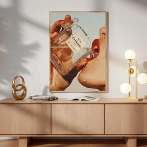 May include: A rectangular wall art print featuring a vintage-style collage of a hand holding a glass Chanel N&deg;5 Eau de Parfum bottle, with the liquid pouring toward a pair of red lips. The artwork is framed in a light wood frame and displayed.
