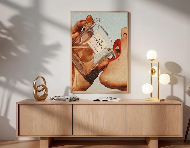 May include: A rectangular wall art print featuring a vintage-style collage of a hand holding a glass Chanel N&deg;5 Eau de Parfum bottle, with the liquid pouring toward a pair of red lips. The artwork is framed in a light wood frame and displayed.