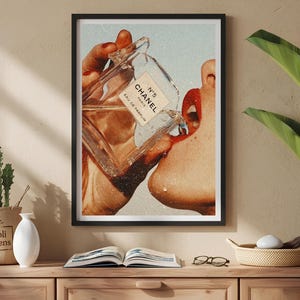 May include: A framed wall art print featuring a vintage-style collage of a hand holding a glass perfume bottle labeled N&deg;5 CHANEL EAU DE PARFUM, with liquid pouring toward a mouth with bright red lipstick, set against a light blue background.