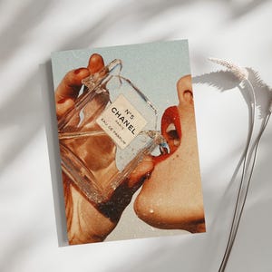 May include: A rectangular art print featuring a vintage-style collage of a clear glass Chanel No. 5 Eau de Parfum bottle being held near a face with bright red lips. The image has a grainy, retro aesthetic with warm, sun-drenched color tones.