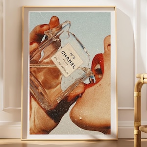 May include: A framed wall art print featuring a collage of a person holding a glass Chanel No. 5 Eau de Parfum bottle, pouring the liquid toward their open mouth. The image displays a vintage aesthetic with red lipstick, red nail polish, and text.