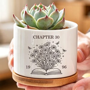Op de afbeelding: Een witte cilindrische keramische plantenpot met een bamboe basis met een groene vetplant. De pot is voorzien van een zwarte lijntekening van een open boek met bloemen, vlinders en sterren, met de tekst "CHAPTER 30" en "19 96".
