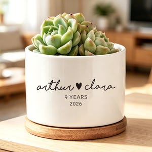 Op de afbeelding: Een witte cilindervormige keramische plantenpot met een groene vetplant. De pot is voorzien van zwarte tekst met "arthur ♥ clara" en daaronder "9 YEARS" en "2026". Het staat op een ronde onderzetter van lichtbruin hout.