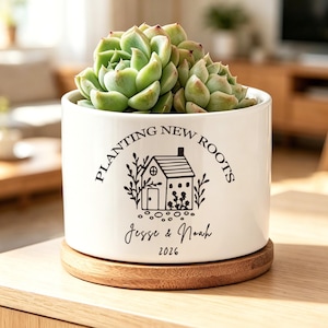 May include: A white ceramic cylindrical planter with a wooden base, containing a green succulent. The planter features black text reading "PLANTING NEW ROOTS," a graphic of a small house, and the names "Jesse & Noah" with the year "2026" below.