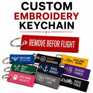 May include: A collection of rectangular embroidered fabric keychains in various colors including red, blue, green, pink, black, yellow, purple, and gray. Each keychain features a metal ring and white text with icons such as REMOVE BEFOR FLIGHT.