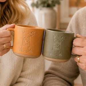 May include: Two ceramic mugs, one mustard yellow and one olive green, each featuring an embossed tree trunk design with a heart containing the letters D.Y. and the date 3.18.25 carved into the center. Both mugs have a smooth, unglazed base.