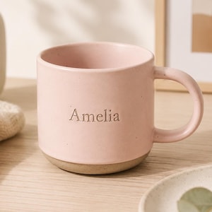 Puede incluir: Una taza de cerámica rosa claro con acabado liso y asa redondeada. El nombre Amelia está grabado en una fuente con serifa en la parte frontal. La base de la taza es de un color beige sin esmaltar que contrasta con el resto de la pieza.