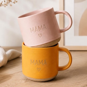 Puede incluir: Una pila de dos tazas de cerámica con acabado mate. La taza superior es de color rosa claro y la inferior es de color amarillo mostaza. Ambas tazas tienen la palabra MAMA y un pequeño corazón grabados en el frente, con una base sin esmaltar.