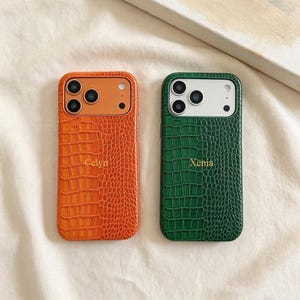 May include: Two rectangular phone cases with a textured crocodile skin pattern. The left case is orange with the name Celyn printed in gold, and the right case is green with the name Xenia printed in gold. Both feature a camera cutout area.