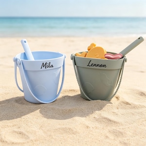 May include: A light blue bucket with the name Mila printed on the side and a sage green bucket with the name Lennon printed on the side. Both buckets feature a handle, a matching shovel, and embossed sea creature shapes on the exterior surface.