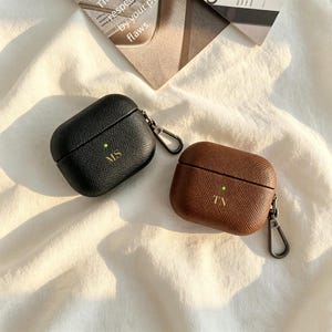 May include: Two textured earbud cases, one black and one brown, each featuring a gold-toned metal carabiner clip on the side. The black case is monogrammed with the letters MS, and the brown case is monogrammed with the letters TA. Both cases display a small, illuminated green LED indicator light on the front.