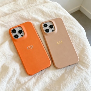 May include: Two smartphone cases with camera cutouts. One case is bright orange with the silver-colored letters GB embossed in the center. The other case is a muted beige color with the gold-colored letters AM embossed in the center.