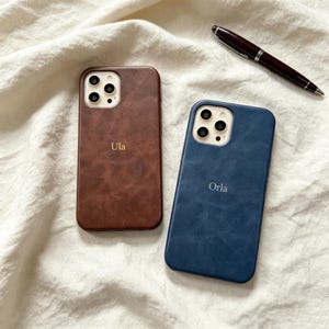 Puede incluir: Dos fundas para smartphone con acabado texturizado, una en color marrón con el nombre Ula grabado en dorado y otra en azul con el nombre Orla grabado en plateado. Un bolígrafo marrón oscuro con detalles plateados se sitúa en la esquina superior.