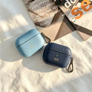 May include: Two textured protective cases for wireless earbuds, one in light blue and one in dark navy blue. Each case features a small metal carabiner clip on the side, a centered green indicator light, and gold-colored monogrammed initials.