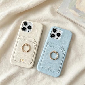 May include: Two textured phone cases, one cream and one light blue, each featuring a card pocket, a gold circular ring stand, and gold embossed initials. The cream case is marked with CK, while the light blue case is marked with RT. Both cases have a triple camera cutout.