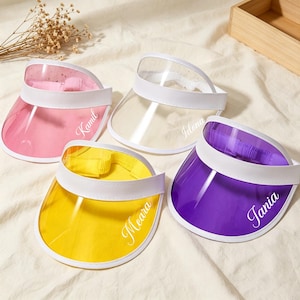 Peut inclure: Quatre visières de soleil en plastique transparent avec bordure blanche et sangle élastique. Les visières sont rose, transparente, jaune et violette. Chaque visière porte un prénom en écriture cursive blanche : Kamil, Idona, Meara et Tania.
