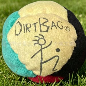 Puede incluir: Una pelota multicolor con el texto "DIRT BAG" y un diseño de figura de palo. La pelota es principalmente amarilla, con acentos turquesa, negros y rojos. La pelota está sobre un lecho de hierba verde.