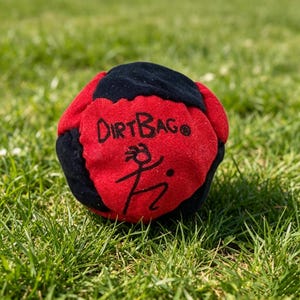 Puede incluir: Una pelota de hacky sack Dirtbag roja y negra con un diseño de figura de palo. La pelota está hecha de tela y está sobre hierba verde. El texto "Dirtbag" está impreso en el panel rojo.