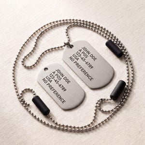 May include: Two silver dog tags with ball chains and black silencers. Each tag has the name "JOHN DOE", address, and phone number. The text "NO PREFERENCE" is also printed on each tag. The tags are displayed on a light-coloured surface.