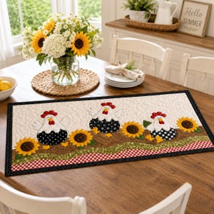May include: A rectangular quilted table runner featuring three white chickens with red combs and black patterned bodies, interspersed with yellow sunflowers on a brown ground, finished with a red and white gingham border and black trim.