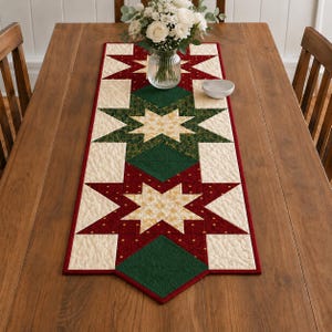 May include: A quilted table runner featuring a repeating star pattern in deep red, forest green, and cream fabric. The design includes gold-toned star centers and a red border. The runner is laid flat on a wooden table with a glass vase of white flowers.