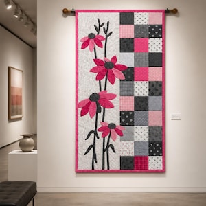 May include: A rectangular wall hanging quilt featuring a vertical split design. The left side displays four pink and dark gray fabric flowers on long stems against a white quilted background. The right side features a checkerboard pattern of pink, gray, and white squares with various small patterns like hearts and dots.
