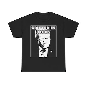 May include: A black short-sleeve t-shirt featuring a high-contrast white graphic print of a person's face in a suit and tie. Above the portrait, the text "GRIFTER IN CHIEF" is printed in bold, white, sans-serif capital letters within a rectangular frame.