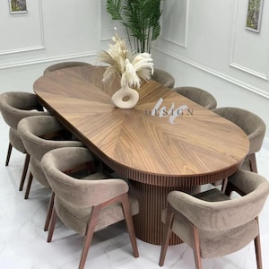 May include: An oval wooden dining table with a sunburst veneer pattern and a fluted cylindrical base, surrounded by eight upholstered chairs with curved backs and wooden frames. A white ceramic vase with dried pampas grass sits on the table.