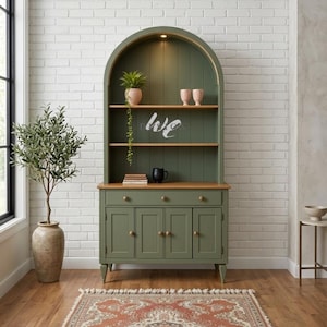 May include: An arched sage green hutch cabinet featuring two open shelves with a light wood finish, three drawers, and four lower cabinet doors with round knobs. The unit includes an integrated top light, a vertical paneled back, and a wooden countertop.