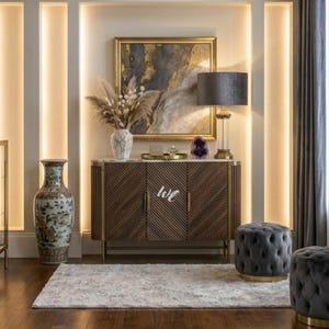 May include: A dark wood sideboard with diagonal slats and gold metal accents, topped with a white marble surface, a white vase with dried pampas grass, a gold tray, a purple amethyst geode, and a table lamp with a dark grey shade.