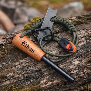 Könnte beinhalten: Ein Feuerstarter mit einem Holzgriff, in den der Name Ethan eingraviert ist, befestigt an einem dunkelolivgrünen Paracord-Lanyard. Das Set enthält einen schwarzen Metall-Schaber mit Lineal und einen kleinen schwarzen Karabiner mit orangefarbener Pfeife.