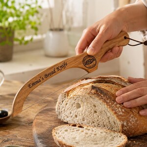 May include: A wooden bread knife with a thin metal wire blade, featuring the engraved text Emma's Bread Knife and a circular logo with a bread illustration. The handle includes a small brown leather cord loop for hanging storage.