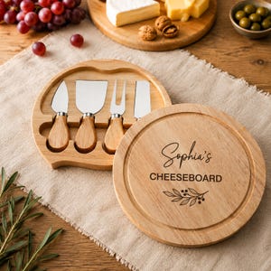 Puede incluir: Una tabla de quesos redonda de madera con tapa giratoria que presenta el texto grabado Sophia's CHEESEBOARD y un diseño de hojas. La tabla contiene cuatro cuchillos de queso de acero inoxidable con mangos de madera clara, incluyendo una pala.